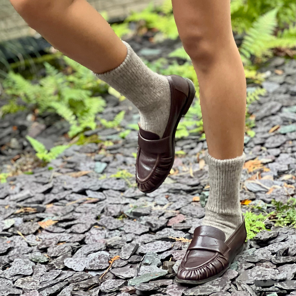 Person wearing brown loafers and light gray  Yak socks on a stone path with greenery.