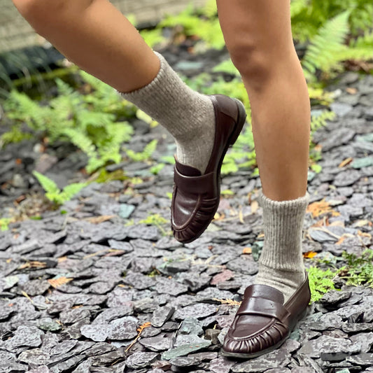 Person wearing brown loafers and light gray  Yak socks on a stone path with greenery.
