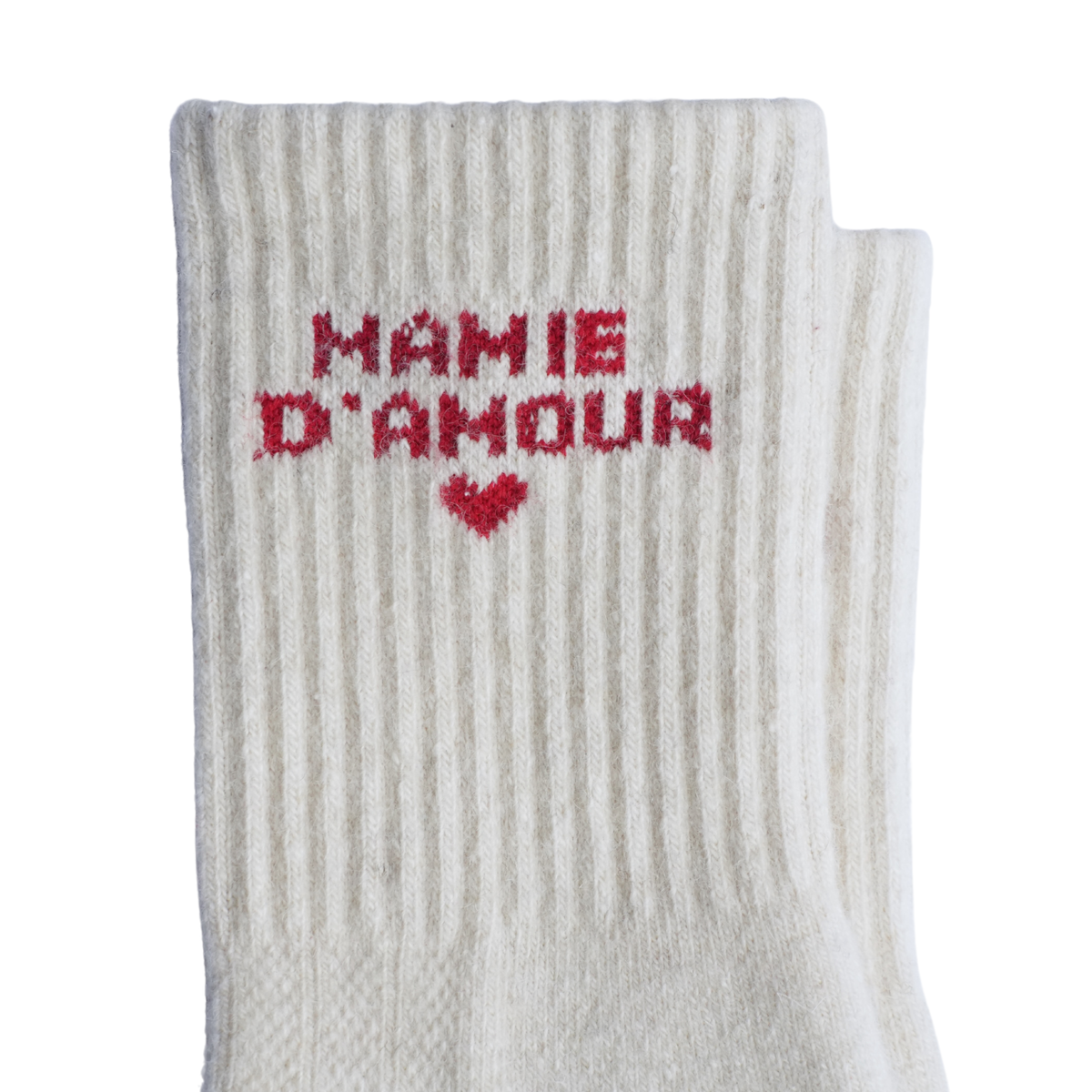 Une chaussette en maille côtelée blanc cassé portant l'inscription « MAMIE D'AMOUR » et un petit cœur tricoté en fil rouge, présentée sur un fond blanc uni.
