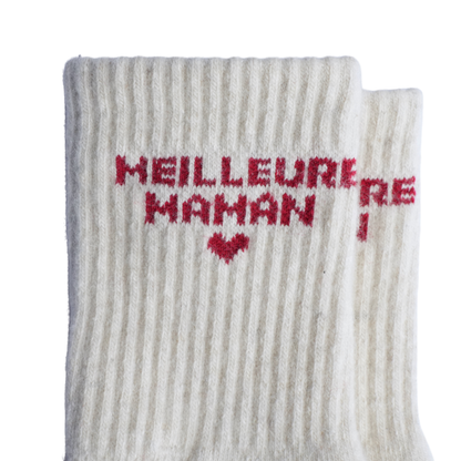 Gros plan sur une chaussette en maille côtelée blanc cassé, ornée de l'inscription « MEILLEURE MAMAN » tricotée en rouge et d'un petit cœur rouge, présentée sur un fond blanc uni.