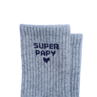Une paire de chaussettes en maille côtelée gris chiné, avec l'inscription « SUPER PAPY » et un cœur tricotés en bleu marine sur le devant, présentées sur un fond blanc uni.
